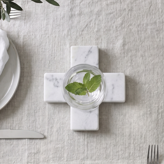 A styled tabletop setting featuring a light beige linen table runner laid across a gray wood-grain table. In the center of the runner sits a white marble cross-shaped coaster holding a small glass of water with green mint leaves.