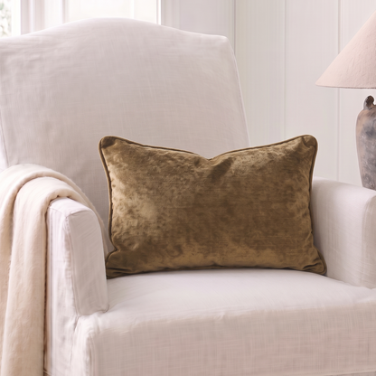 A square olive-brown velvet throw pillow resting on a white upholstered armchair in a bright, neutral room setting with a lamp on a side table.