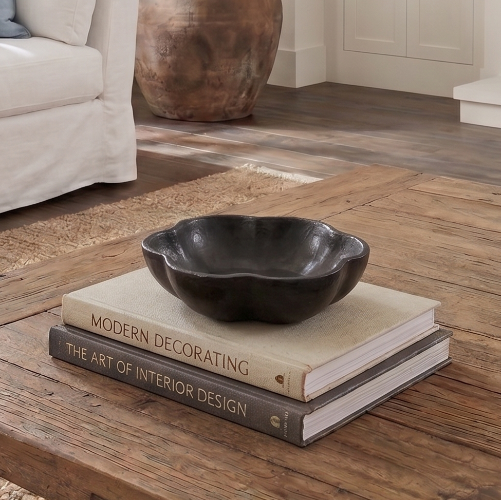 A dark charcoal black scalloped ceramic bowl sits on top of two decorative books on a rustic wooden coffee table in a styled living room setting.