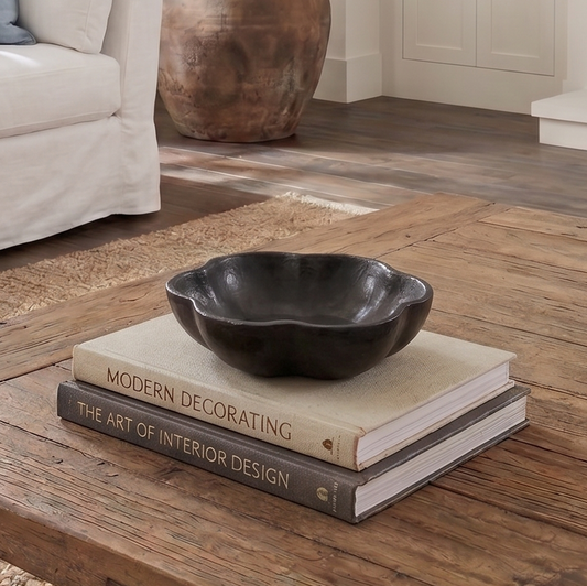 A dark charcoal black scalloped ceramic bowl sits on top of two decorative books on a rustic wooden coffee table in a styled living room setting.