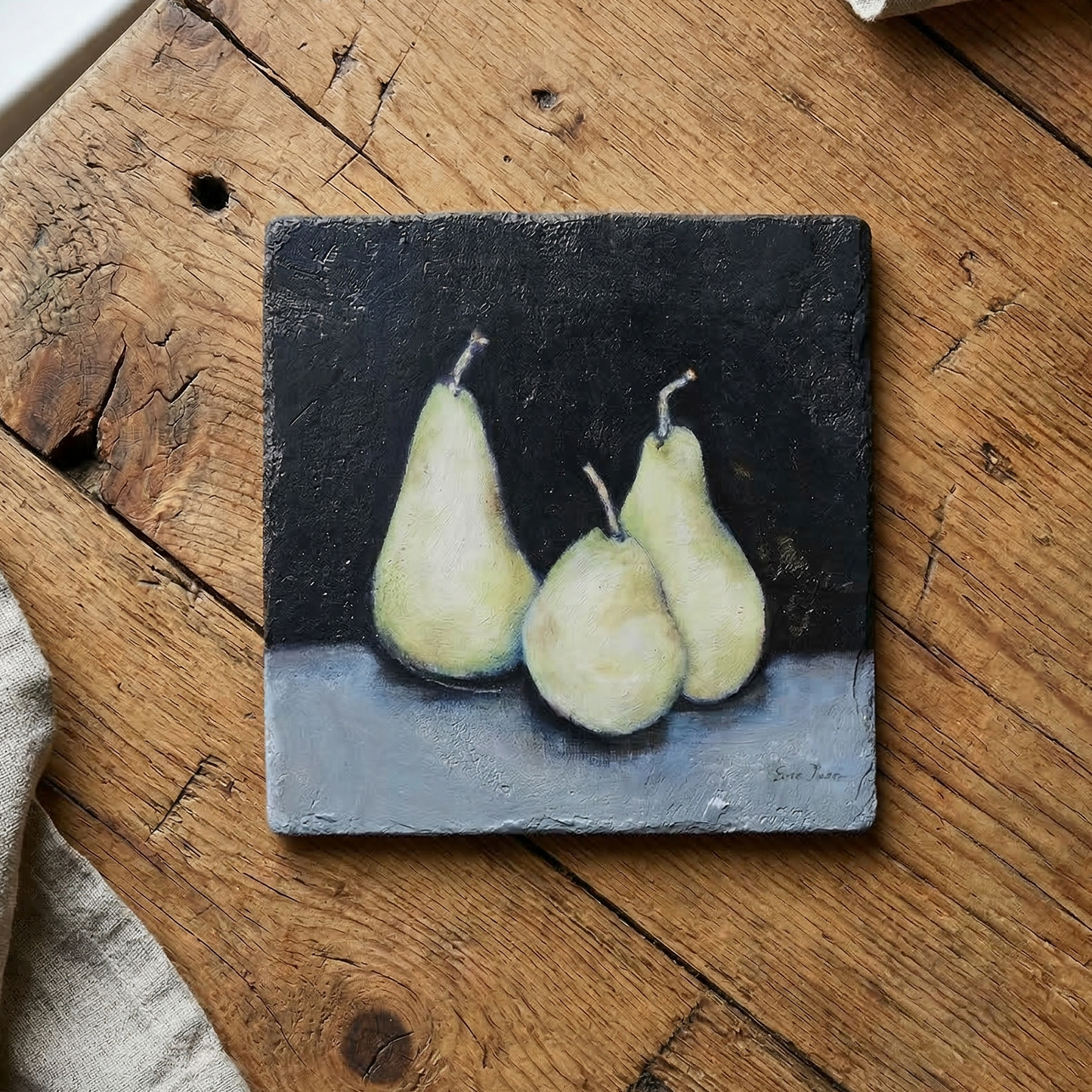 Small painting of three pears on a trivet sitting on a wooden surface with a cloth and small containers.