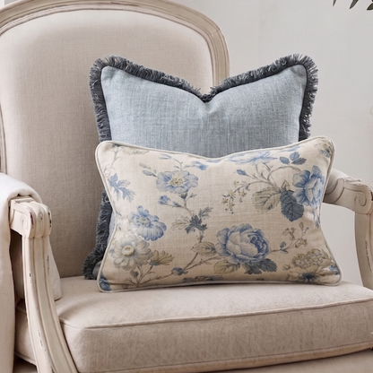 Two decorative throw pillows displayed on a distressed cream-colored armchair. The front pillow is a rectangular lumbar cushion with a beige linen base and a blue floral pattern, while the back pillow is a square cushion in a dusty blue linen fabric with a dark blue fringe.