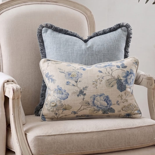 Two decorative throw pillows displayed on a distressed cream-colored armchair. The front pillow is a rectangular lumbar cushion with a beige linen base and a blue floral pattern, while the back pillow is a square cushion in a dusty blue linen fabric with a dark blue fringe.