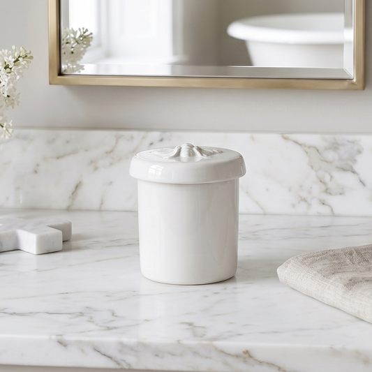 A white ceramic lidded container with a decorative embossed top sits on a white marble bathroom countertop, with a matching marble backsplash, a folded beige cloth, and a mirror in the background.