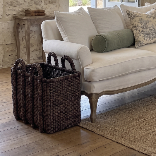 A cream-colored upholstered loveseat with patterned and solid throw pillows, styled in a rustic stone-walled living room with a fireplace, woven baskets, and a jute rug.