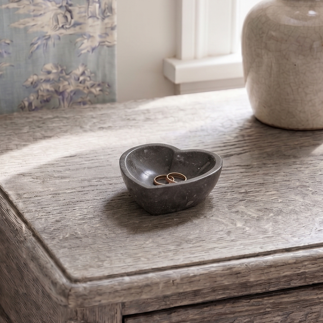 A small heart-shaped dark gray stone ring dish holding two gold rings, placed on a rustic gray wooden table next to a beige ceramic table lamp.
