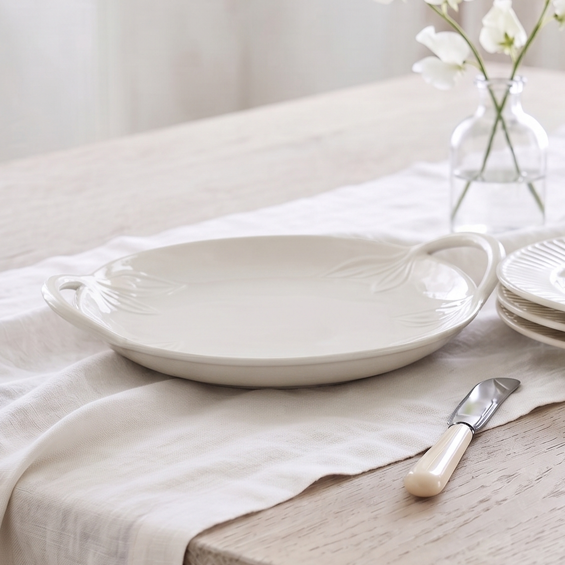 A styled dining table setting featuring a white ceramic serving platter with handles, a stack of matching plates, a small glass vase with white flowers, and a butter knife on a light wood table covered with a cream linen runner.