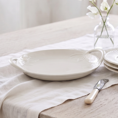 A styled dining table setting featuring a white ceramic serving platter with handles, a stack of matching plates, a small glass vase with white flowers, and a butter knife on a light wood table covered with a cream linen runner.