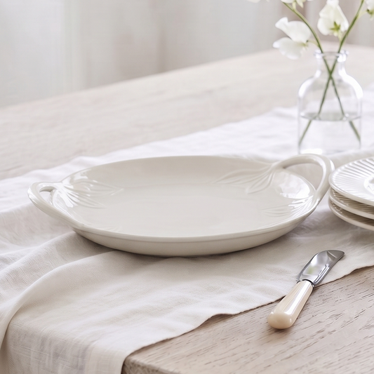 A styled dining table setting featuring a white ceramic serving platter with handles, a stack of matching plates, a small glass vase with white flowers, and a butter knife on a light wood table covered with a cream linen runner.