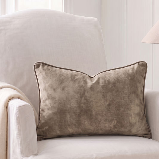 A rectangular throw pillow in a textured taupe velvet fabric with piped edges, resting on a white upholstered armchair in a bright, neutral-toned room.
