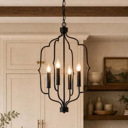 A black metal five-light chandelier with a decorative open frame hangs in a warm, rustic kitchen setting with cream cabinets and wooden beams.