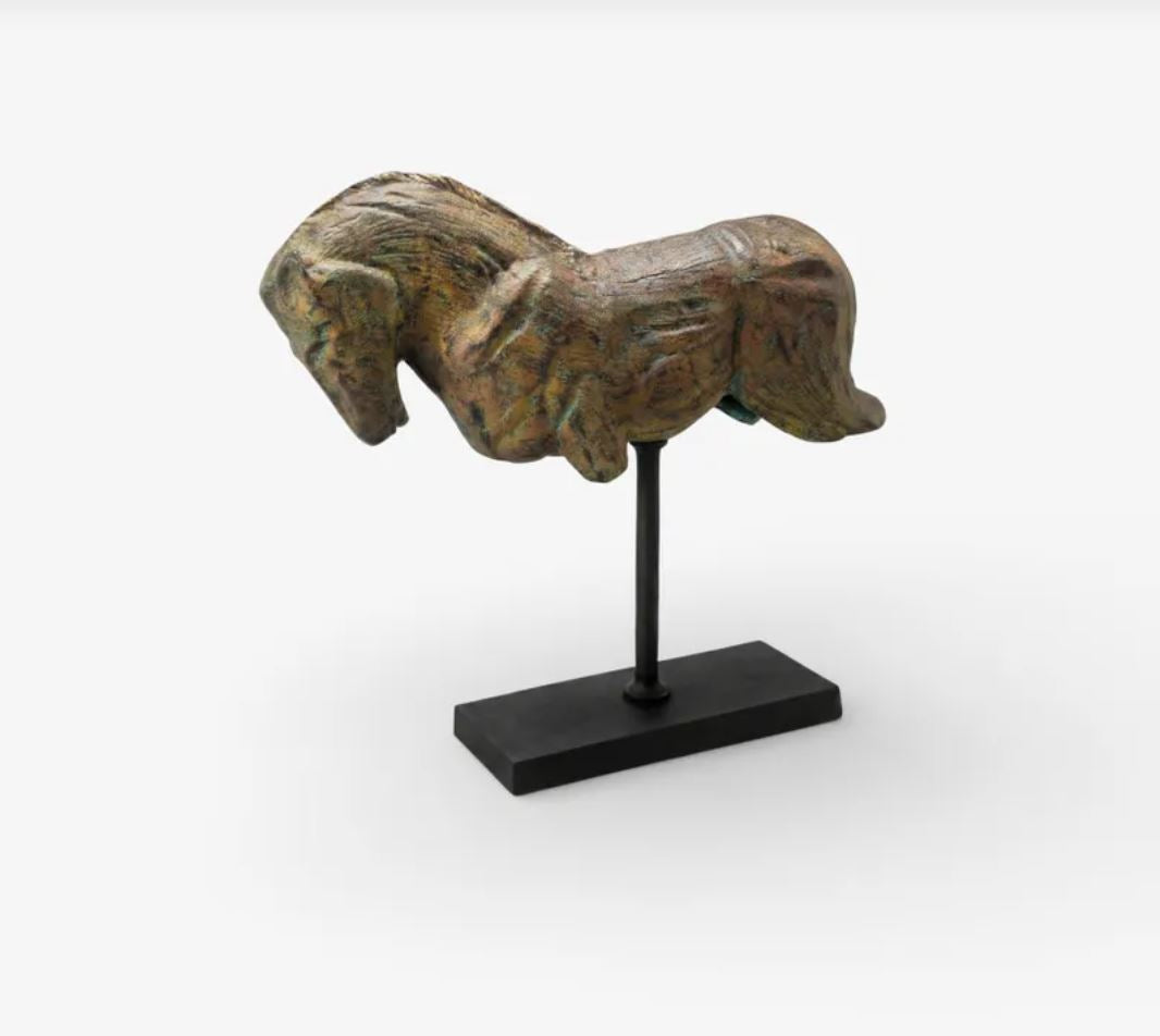 A small sculptural horse figurine with a textured, weathered bronze-brown finish mounted on a black display stand against a light gray background.