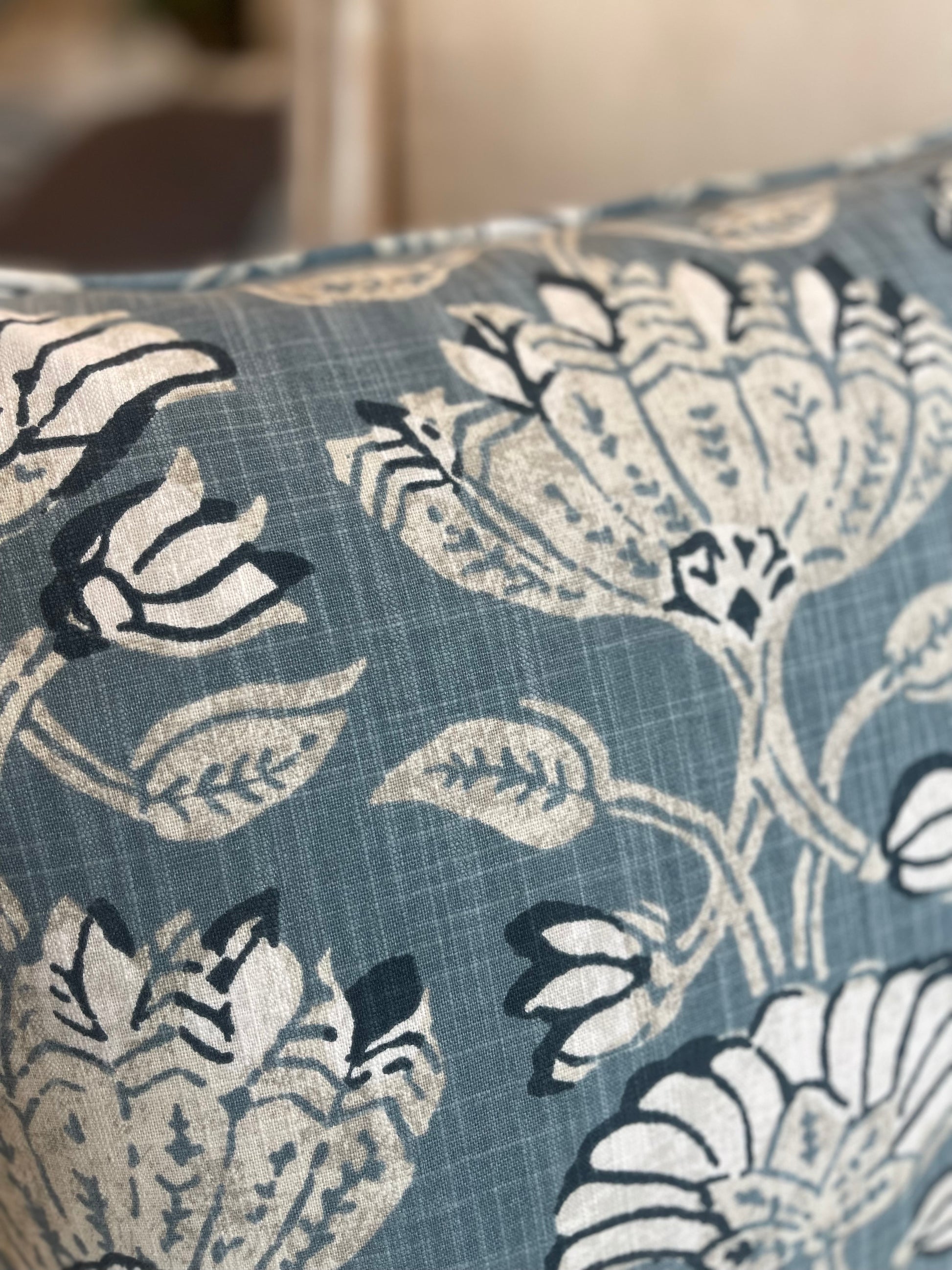 The heavyweight Pradesh Art Deco Floral Blue Taupe Cushion, featuring a refined pattern from Robert Allen Fabrics, is sure to add a touch of sophistication to any room. Utilizing a composition of pale taupe, off white, and blue hues, its feather-filled insert provides unparalleled comfort for a truly cozy feel. Close up.