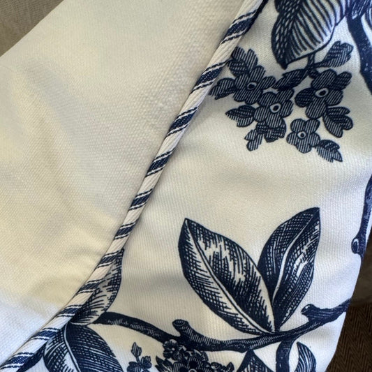 A close-up of a decorative throw pillow featuring a navy blue floral and leaf pattern on an off-white background, resting on a beige upholstered chair.
