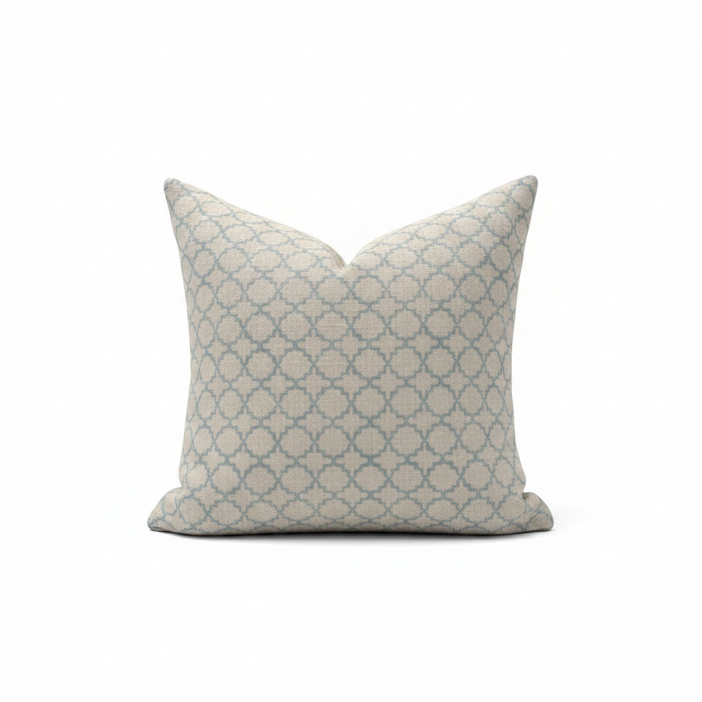 A square throw pillow with a blue and beige geometric trellis pattern, resting on a light beige upholstered armchair.