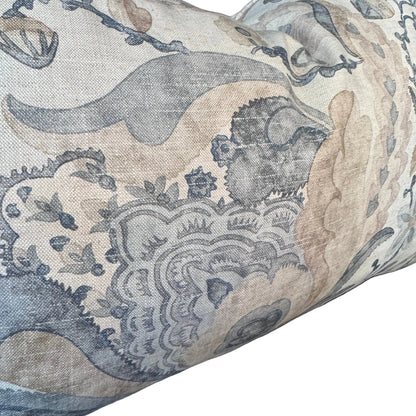 A close-up of a decorative throw pillow featuring an intricate botanical and scroll pattern in muted blue-gray and beige tones on a textured fabric, set against a dark background.