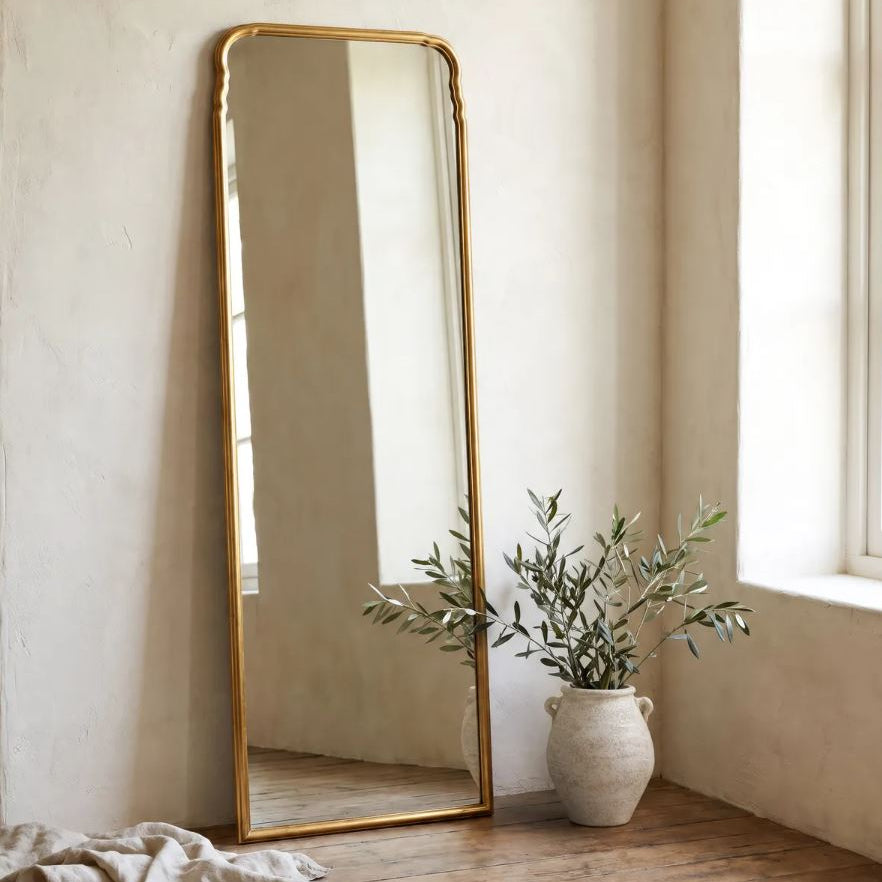 A tall floor mirror with a gold frame leans against a textured cream wall on a rustic wooden floor, next to a white ceramic vase with green branches and a draped beige cloth.