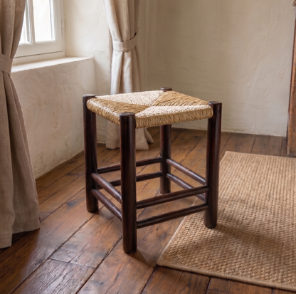 Wooden stool with woven seat on a wooden floor in a room with curtains and a rug.