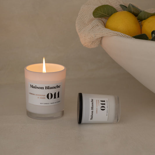 Candle with a label and a small container next to a bowl of lemons on a neutral background