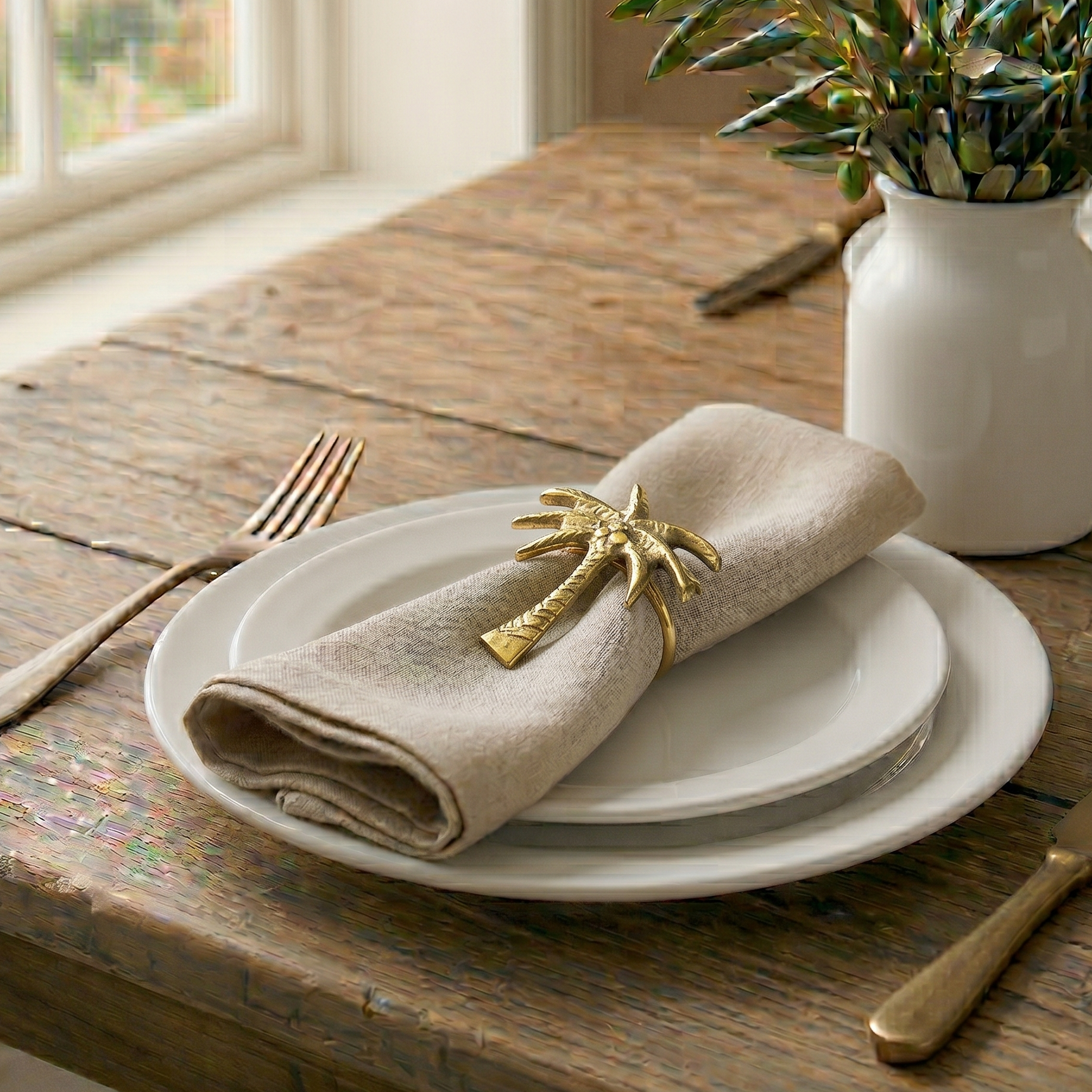Table setting with folded beige napkin, gold palm tree napkin ring, and white plates on a wooden table.