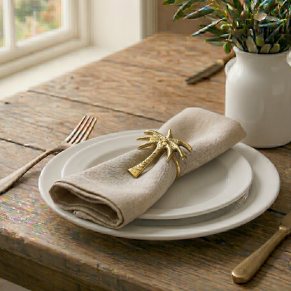 Table setting with folded beige napkin, gold palm tree napkin ring, and white plates on a wooden table.