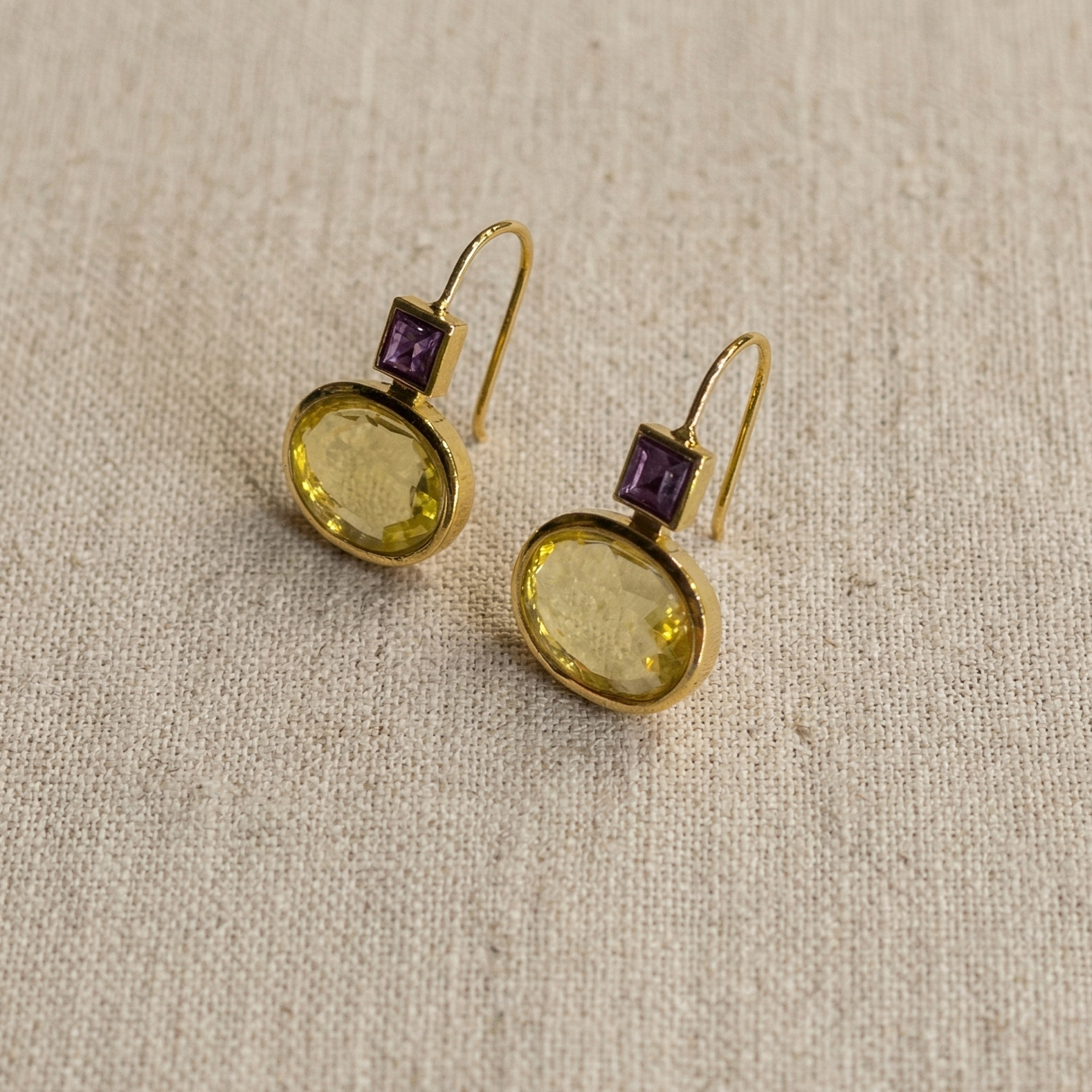 Gold earrings with purple and green gemstones on a beige fabric background