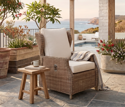 Outdoor patio with wicker chair, cushions, and a small table with a cup, set against a scenic backdrop.