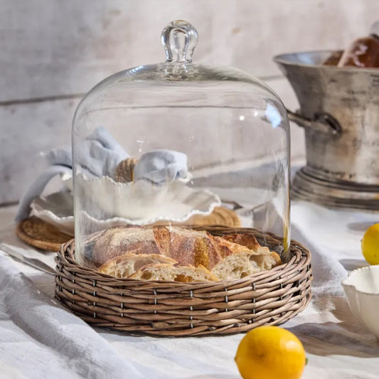 Glass cloche over a basket of bread on a table with lemons and a bowl.
