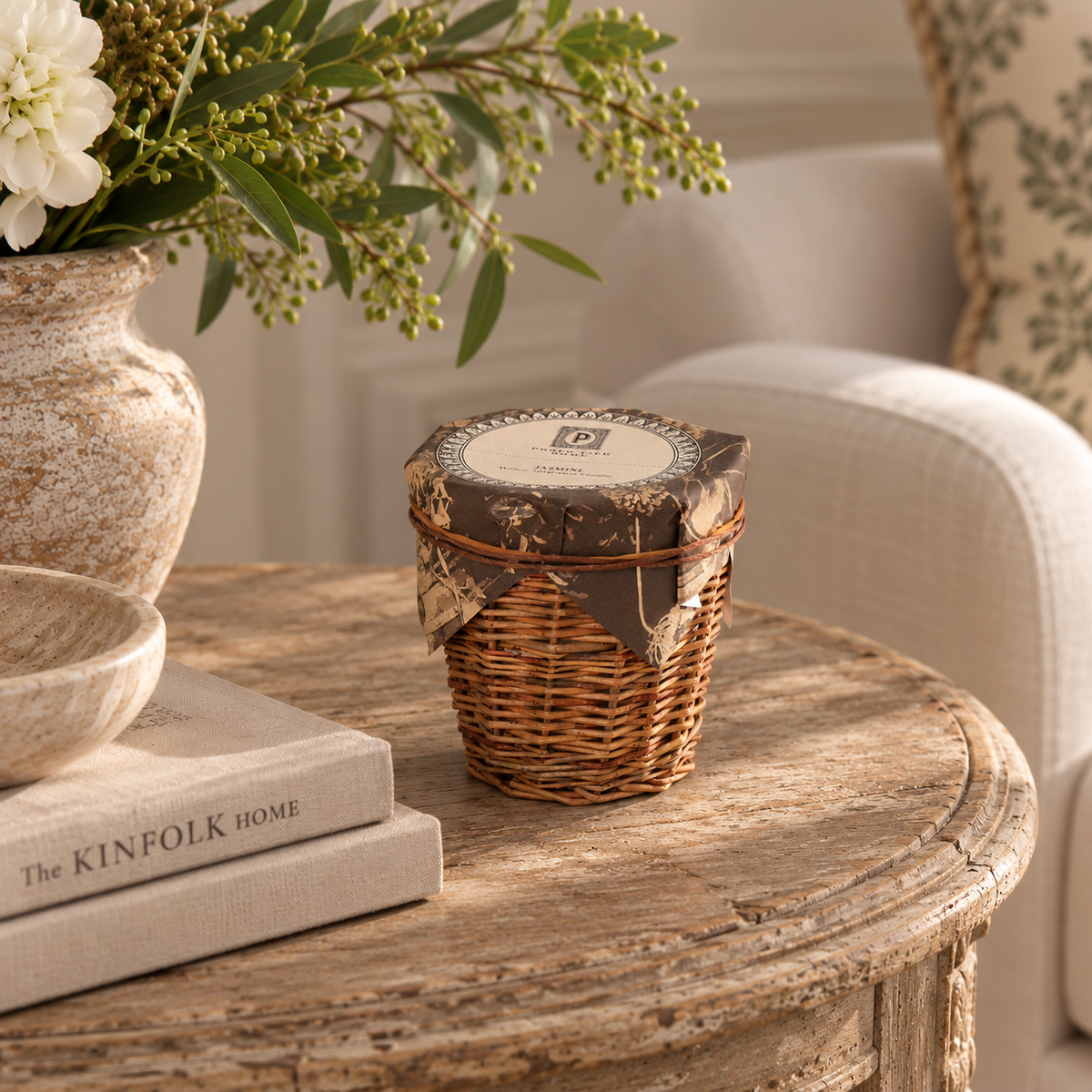 A small wicker basket jar with a brown patterned lid wrap sits on a rustic wooden table in a styled living room setting next to a vase of white flowers and books.
