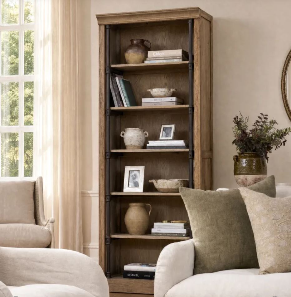 A tall rustic wooden bookcase with black metal frame accents, styled in a cozy living room next to a window and sofa.