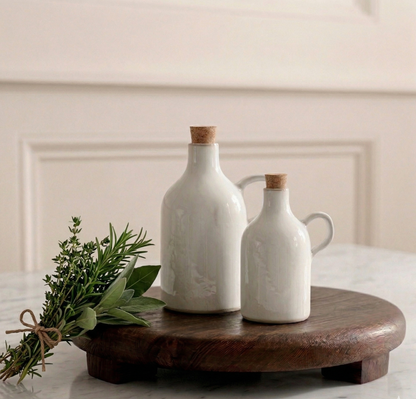 Two white ceramic bottles with wooden lids on a wooden stand with greenery against a beige wall.