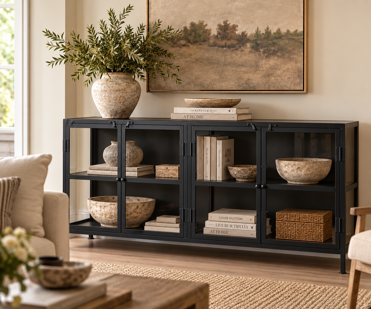 A black metal sideboard cabinet with glass doors, styled in a cozy living room setting with books, decorative bowls, a large ceramic vase with greenery, and a framed landscape painting on the wall.