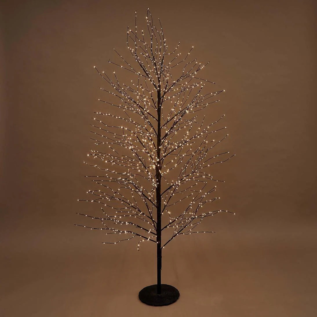 A bare artificial tree with a black trunk and branches, decorated with many small warm white LED lights, standing on a round black base against a plain brown studio background.