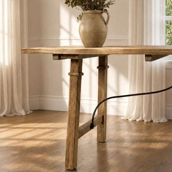 A rustic wooden table with a thick rectangular top and trestle-style legs, shown in a sunlit room with cream curtains, a wood floor, and a ceramic vase on top.