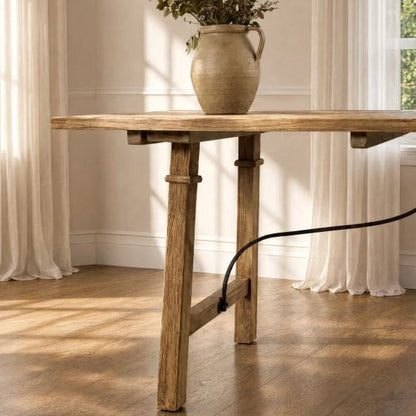 A rustic wooden table with a thick rectangular top and trestle-style legs, shown in a sunlit room with cream curtains, a wood floor, and a ceramic vase on top.