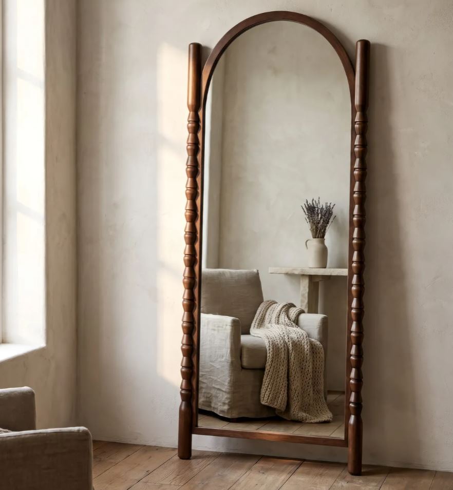A tall arched full-length floor mirror with a dark brown wooden frame and turned corner posts, standing in a softly lit room with a beige armchair and small side table reflected in the glass.