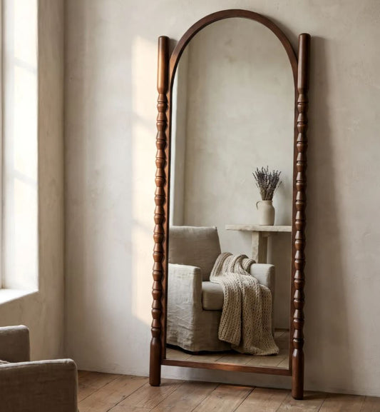 A tall arched full-length floor mirror with a dark brown wooden frame and turned corner posts, standing in a softly lit room with a beige armchair and small side table reflected in the glass.