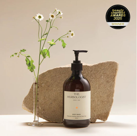 Bottle of 'The Herbologist' body wash with a rock and flowers on a beige background, featuring an award badge.