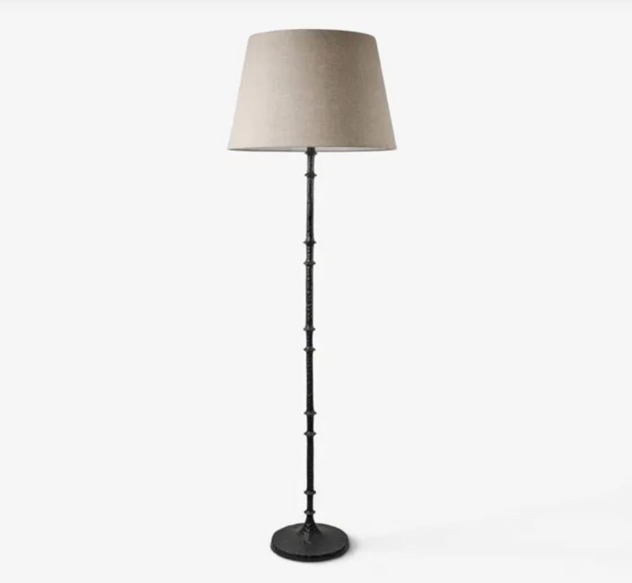 A floor lamp with a tapered beige fabric drum shade and a dark metal stem with decorative ring details, standing on a round base against a light gray background.