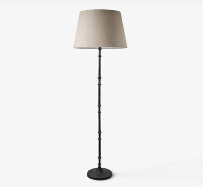 A floor lamp with a tapered beige fabric drum shade and a dark metal stem with decorative ring details, standing on a round base against a light gray background.