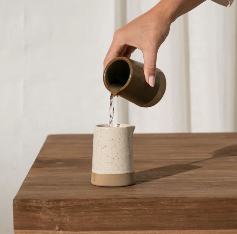 Person pouring water from a ceramic pitcher into another on a wooden surface.