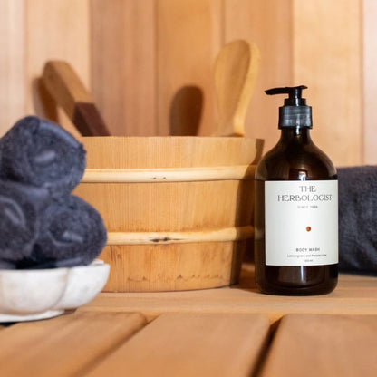 Bottle of 'The Herbologist' body wash on a wooden surface with towels and a wooden basket in the background.