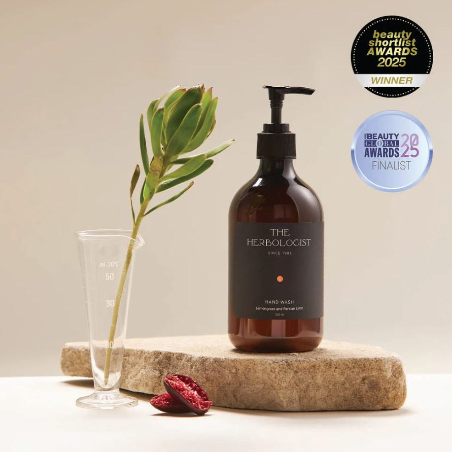 Brown bottle labeled 'The Herbologist' on a stone surface with a plant and pomegranate seeds, against a beige background.