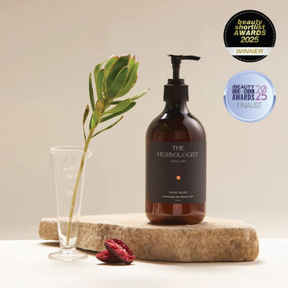 Brown bottle labeled 'The Herbologist' on a stone surface with a plant and pomegranate seeds, against a beige background.