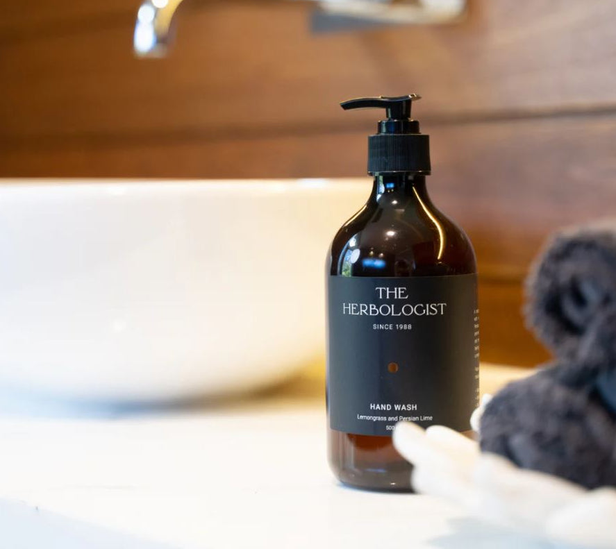 Bottle of 'The Herbologist' hand wash on a bathroom counter with a sink and towel in the background.