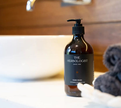 Bottle of 'The Herbologist' hand wash on a bathroom counter with a sink and towel in the background.