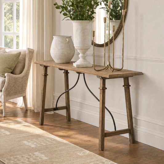 A rustic wooden console table in a bright living room setting, styled with a large white urn-shaped planter, a smaller textured vase, and a gold candlestick holder, with a round mirror and window in the background.