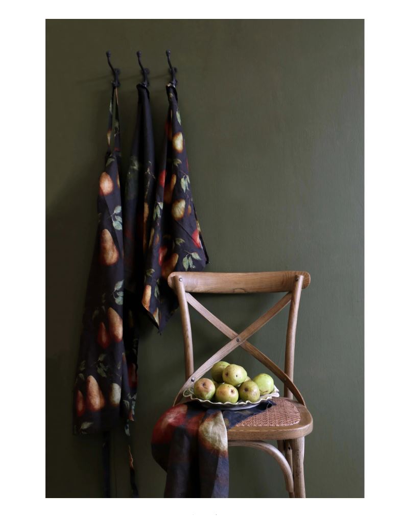 Three patterned cloths hanging on hooks against a green wall with a wooden chair and fruit bowl.