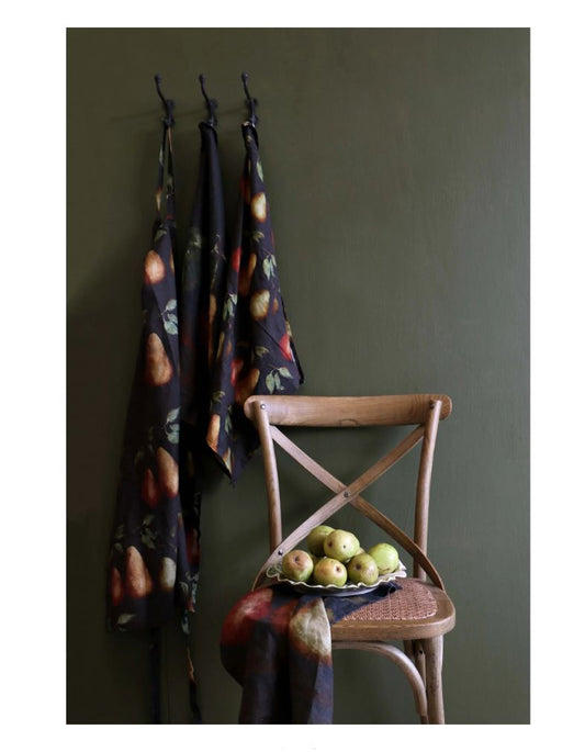 Three patterned cloths hanging on hooks against a green wall with a wooden chair and fruit bowl.