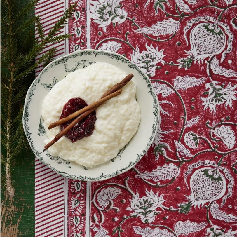 A top-down view of a white plate with a green floral border, filled with creamy white pudding or custard and topped with a dark red jam dollop and two cinnamon sticks, set on a red and white patterned tablecloth with evergreen branches.